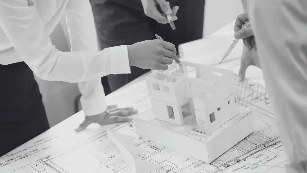 Architekci omawiają projekt domu, analizując model i plany. Kara za podniesienie ścianki kolankowej może wpłynąć na ostateczny kształt budynku.